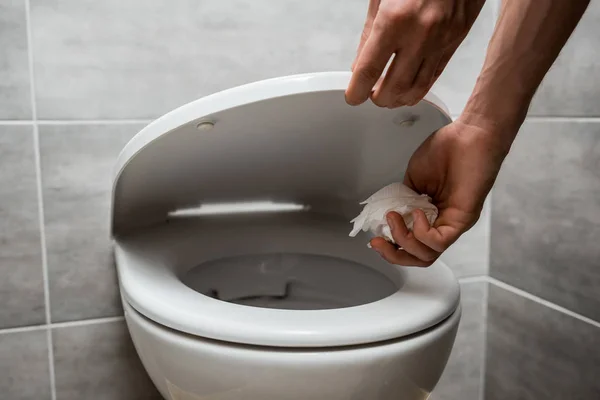 Cropped view of man holding toilet lid and throwing paper in toilet bowl