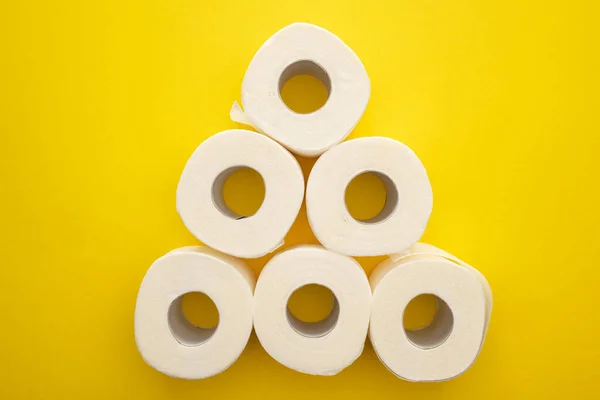 Top view of white toilet paper rolls arranged in pyramid on yellow background